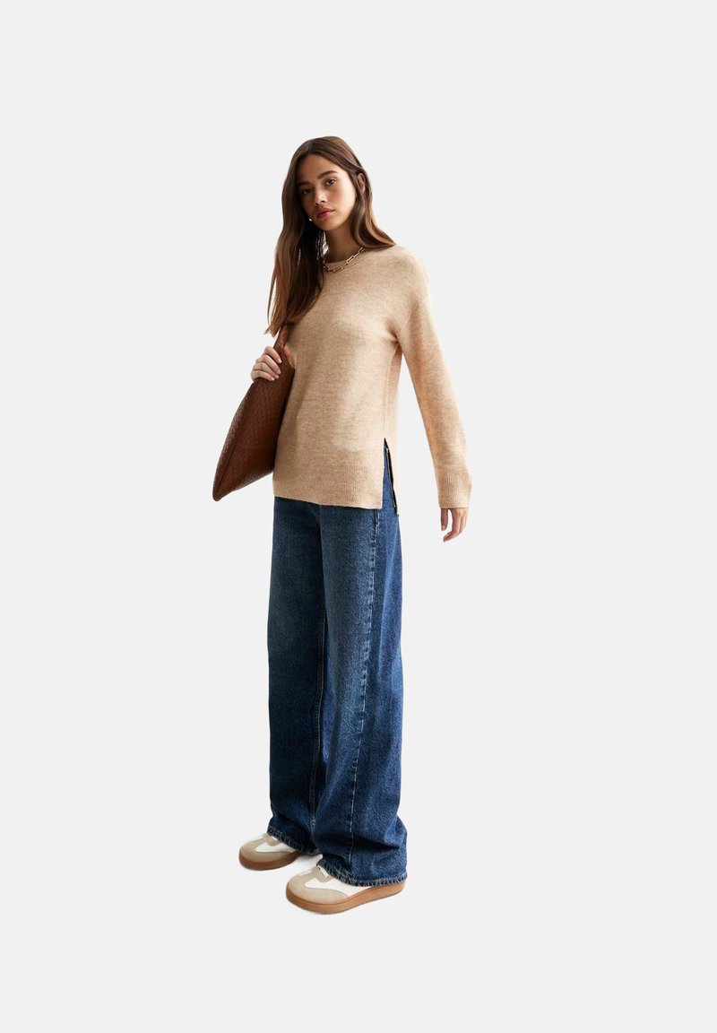 Maglione beige lavorato a maglia con fessure laterali, abbinato a jeans a gamba larga blu e sneakers chiare. La modella tiene una borsa a tracolla marrone con motivo.