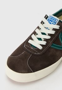 Barna velúr sneaker fehér fűzőkkel és varrással. Zöld oldali logóval rendelkezik. Kerek orrú és krémszínű gumi talpa van a tapadás érdekében.