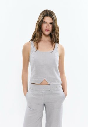 Jeune femme aux longs cheveux ondulés portant un haut sans manches gris clair et un pantalon assorti, les mains dans les poches, debout devant un fond blanc.