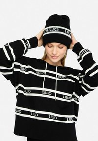 Felpa nera corta con strisce orizzontali bianche e logo "LIUJO". Abbinata a un berretto a maglia nero con dettaglio a strisce bianche.