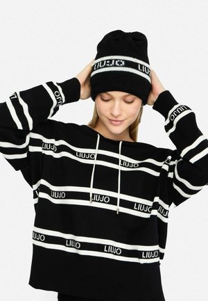 Felpa nera corta con strisce orizzontali bianche e logo "LIUJO". Abbinata a un berretto a maglia nero con dettaglio a strisce bianche.