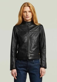 Donna con lunghe capelli rossi che indossa una giacca in pelle nera con zip e jeans blu scuro, in piedi contro uno sfondo chiaro e semplice.