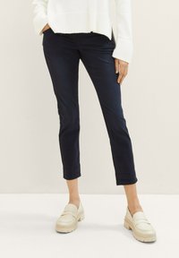 Tmavě modré slim-fit kalhoty ankle length. Vyrobeno z měkké tkaniny, s bočními kapsami. V kombinaci se světlými robustními mokasíny.
