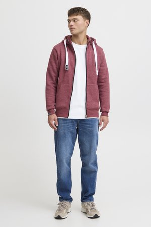 Rode hoodie met witte trekkoorden, rits aan de voorkant en twee zakken, gecombineerd met een witte t-shirt, blauwe spijkerbroek en beige sneakers.