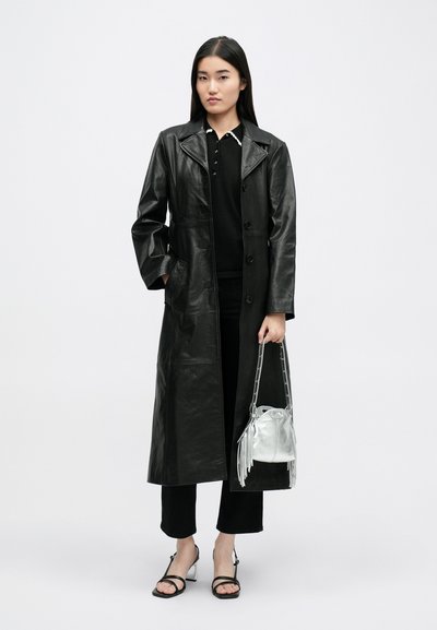 Lungo cappotto di pelle nera con colletto a punte, chiusura con bottoni e tasche laterali. Abbinato a un top nero, pantaloni e una borsa a frange argento.