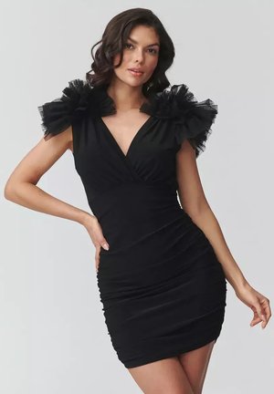 Kobieta w dopasowanej czarnej sukience z marszczonymi tiulowymi ramionami, pozująca z jedną ręką na biodrze na gładkim jasnym tle.