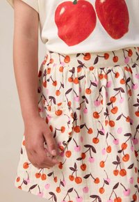 Jupe en coton avec un design évasé, fond crème et motifs de cerises vifs en orange et rose. Elle dispose d'une taille froncée et d'une poche.