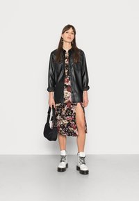 Camisa preta de imitação de couro com botões sobre um vestido florido com uma fenda lateral, combinada com botas de cano curto brancas com atacadores e uma bolsa preta.