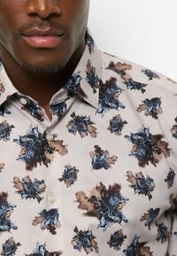 Patterned shirt in light beige with floral designs in blue and brown. Features a classic collar and buttons, crafted from woven fabric.