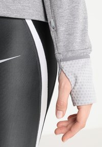 Maglia grigia a maniche lunghe con polsini testurizzati e foro per il pollice, abbinata a leggings neri con strisce bianche laterali e logo.