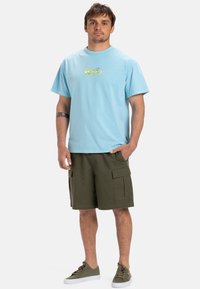 Light blue short-sleeve T-shirt with a yellow graphic design on the chest, paired with olive green cargo shorts and green sneakers.