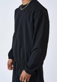 Schwarzes Sportoberteil mit Rundhalsausschnitt und langen Ärmeln, ausgestattet mit elastischen Bündchen und einem Kordelzugsaum. Dezenter strukturiertes Material.