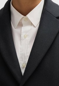 Blazer di lana nera con colletto asciutto, indossato sopra una camicia bianca con bottoni chiari e una texture liscia.