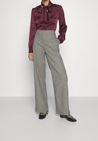 Blusa di raso bordeaux con fiocco, abbinata a pantaloni a gamba larga a scacchi grigio chiaro e neri, indossata con scarpe basse nere.