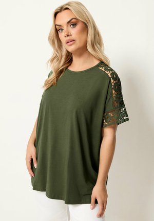Lyshåret kvinde iført løs olivengrøn top med blomstrede blondeudskæringer på ærmerne, parret med hvide bukser, stående mod en ensfarvet baggrund.