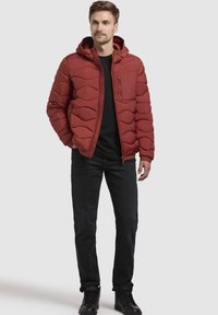 Röd puffjacka med huva, med kviltad textur och en dragkedjeförsedd bröstficka. Bärs över en svart skjorta, i kombination med mörka jeans och svarta skor.
