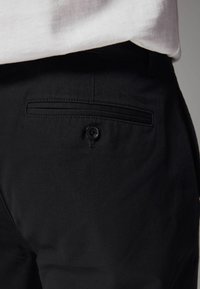 Pantalons noirs à texture lisse, dotés d'une poche arrière avec une fermeture à bouton et de délics coutures décoratives le long du bord de la poche.