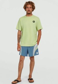 Ανοιχτό πράσινο βαμβακερό μπλουζάκι με στρογγυλό λογότυπο, συνδυασμένο με μπλε σορτς που έχουν καμπύλες λευκές ρίγες και μια κυκλική στάμπα. Μαύρες σανδάλες.