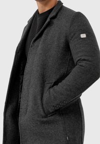 Grau melierter Wollmantel mit Reverskragen, zwei Fronttaschen und geknöpften Manschetten. Verfügt über eine gesteppte Innenlage für zusätzliche Wärme.