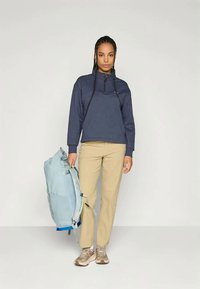 Pulover din fleece bleumarin cu fermoar la jumătate, pantaloni bej și tenisi bej. Poartă un rucsac albastru deschis cu accente albastre închise.