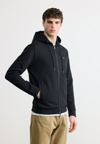 Jeune homme portant un sweat à capuche noir zippé, les mains dans les poches, debout devant un fond clair uni.