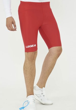 Gambe maschili che indossano pantaloncini da ciclismo rossi aderenti con il logo "LEGEA" e sneakers bianche su sfondo bianco uniforme.