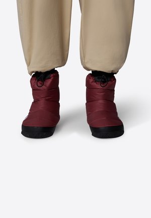 Person iført beige bukser og bordeaux isolerede ankelstøvler med sorte såler, stående på en lys baggrund.