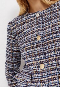 Giacca in tweed con un motivo blu, navy e crema, con bottoni dorati e una finitura texture. Collo rotondo e vestibilità su misura.