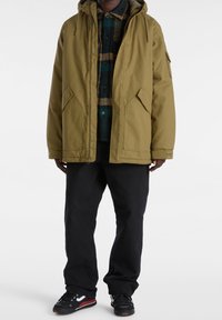 Olive green parka with a hood, two front pockets, worn over a checkered shirt. Black pants and sporty shoes complete the outfit.