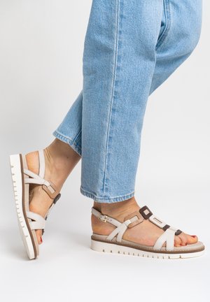 Piedi che indossano sandali bianchi piatti con dettagli in fibbia marrone e jeans azzurri su sfondo bianco semplice.