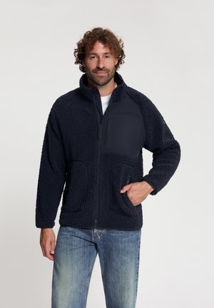 Man met krullend haar die een marineblauw fleecejack met ritssluiting aan de voorkant en zakken draagt, gecombineerd met lichtblauwe jeans, staande tegen een witte achtergrond.