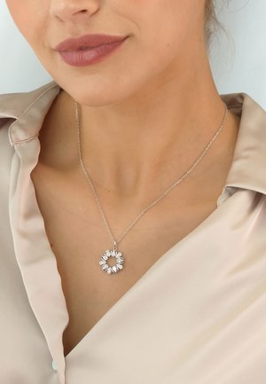 Silberne Halskette mit floralem Design, versehen mit birnenförmigen Akzenten und einer dünnen Kette, präsentiert vor einer hellen Bluse.
