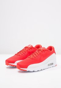 Une paire de baskets Nike Air Max rouges vives avec des semelles intermédiaires blanches, des tiges perforées et des unités de coussinage d'air visibles, présentées sur une surface blanche.