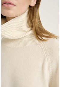 Kobieta w kremowym swetrze z prążkowanym golfem, z prostymi jasnobrązowymi włosami opadającymi na ramie.