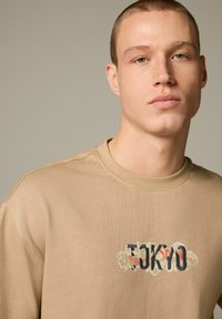 Camiseta beige de manga corta, con un estampado gráfico de "TOKYO" en azul y rojo, con un diseño texturizado en el centro.