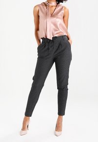 Satinblus i rosa med knytkrage och ärmlös design, i kombination med högmidjade svarta rutiga byxor och beige högklackade skor.