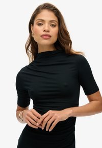 Haut noir à manches courtes avec un col montant, présentant une texture lisse et une coupe ajustée. Mise en valeur par un subtil détail de drapée.