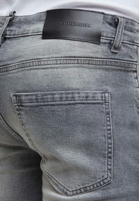 Közelkép a hátsó zsebről és a fekete bőr márkajelzésről, amely egy világosszürke farmernadrágon található, amit egy személy visel.