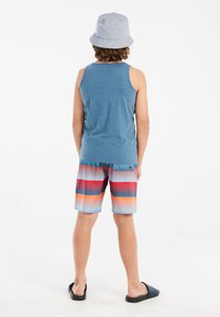 Blauwe tanktop, multicolor gestreepte zwembroek met rood, oranje en blauw. Zwarte instapsandalen en een lichtgrijze bucket hat.