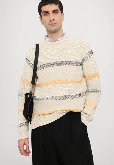 Pull en tricot crème avec des rayures tressées noires et orange, décolleté large et poignets côtelés, associé à un pantalon noir et un petit sac.