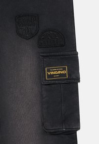 Zwarte cargobroek met geweven patches waarop "CAMP VINGINO" en "EXPLORE" staat. Bevat een stoffen label met "VINGINO" in het geel.