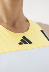 Camiseta deportiva amarilla y blanca sin mangas con escote redondo, que presenta un logo negro de Adidas y acentos negros en los lados; textura suave.