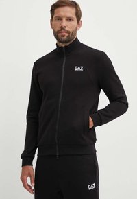 Felpa nera con zip e colletto alto, design aderente e tasche laterali. Presenta un logo EA7 bianco sul petto sinistro. Tessuto liscio.