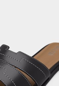 Sandale en cuir noir à double brides avec surpiqûres blanches et semelle intérieure beige avec le nom de la marque partiellement visible.