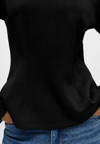 Melnas piegļautās t-krekls ar īsām piedurknēm, izgatavots no gludas auduma, ar šuvju detaļām un viegli noapaļotu apakšmalu. Apvienots ar zilām džinsu biksēm.