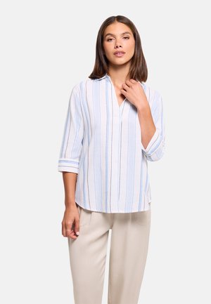 Mujer con pantalones beige y camisa blanca abotonada con rayas verticales azules, posando con una mano cerca del cuello.