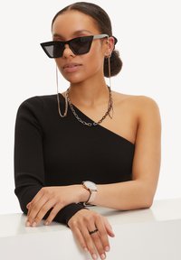 Schwarzes, geripptes Top mit einem Schulterträger, eckige schwarze Sonnenbrille, silberne Kettelemente, hellfarbige Uhr mit rundem Ziffernblatt, zartem Ring am Finger.