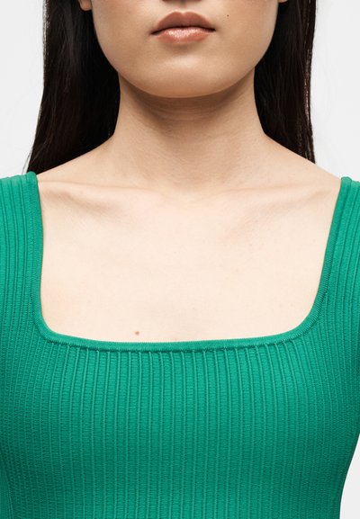 Gros plan d'une personne portant un haut sans manches côtelé vert avec un décolleté carré, montrant le cou, les clavicules et les lèvres.