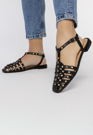 Sandalias de cuero negro con acentos de tachuelas y un diseño de rejilla. Presentan una correa en el tobillo con hebilla y una suela de perfil bajo. Diseño plano y destalonado.