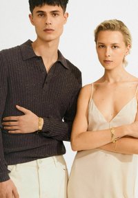 Jonge man in een donker geribbeld sweater en lichte broek en een vrouw in een beige mouwloze jurk, beiden met gouden schakelarmbanden, staan dicht bij elkaar.
